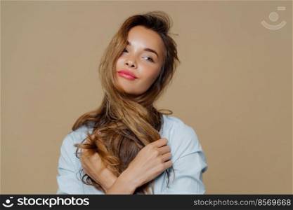 Portrait of thoughtful beautiful woman looks aside, thinks about something pleasant, crosses hair over neck, wears makeup, stylish shirt, isolated over brown background. People, beauty, lifestyle