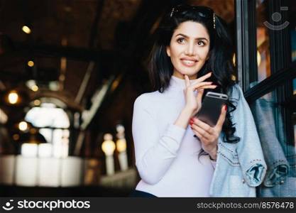 Portrait of thoughtful glamour woman with dark hair wearing sunglasses and white formal clothes surfing social networks using smartphone enjoying online communication posing against cafe interior