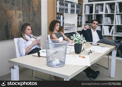 Portrait of three colleagues are boring at table in office and businessman throwing paper in basket. Colleagues boring in office and businessman throwing paper on basket