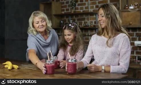 Portrait of three generation family toasting red berry smoothies in mason jars and smiling over modern kitchen background. Beautiful grandmother, daughter and little grandaughter holding fresh berry smoothie and looking at cam with toothy smiles.