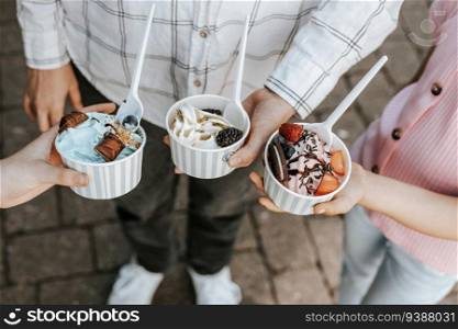 Portrait of three unrecognizable girls holding paper cups with ice cream, berries and cookies standing on a city street on a clear sunny day,. Portrait of three girls with ice cream in their hands.