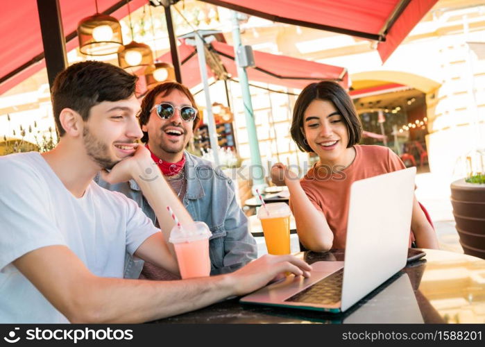 Portrait of three young friends using a laptop while sitting outdoors at coffee shop. Friendship and technology concept.