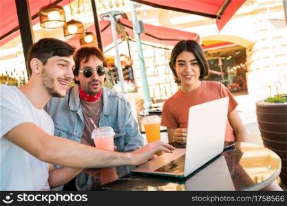 Portrait of three young friends using a laptop while sitting outdoors at coffee shop. Friendship and technology concept.