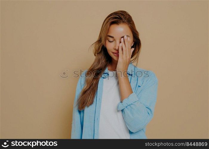 Portrait of tired and annoyed young female hiding eyes as being embarrassed making facepalm gesture tilting head down standing upset and irritated over beige background in blue oversized shirt. Portrait of tired and annoyed young woman posing in studio