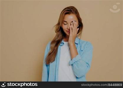 Portrait of tired and annoyed young female hiding eyes as being embarrassed making facepalm gesture tilting head down standing upset and irritated over beige background in blue oversized shirt. Portrait of tired and annoyed young woman posing in studio