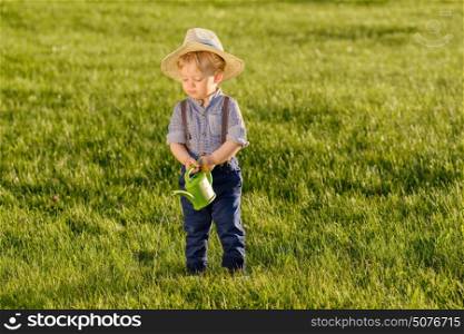 Portrait of toddler child outdoors. Rural scene with one year old baby boy wearing straw hat using watering can