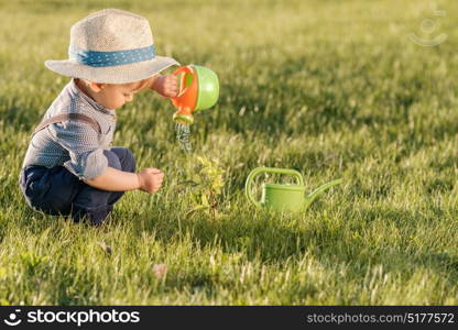 Portrait of toddler child outdoors. Rural scene with one year old baby boy wearing straw hat using watering can