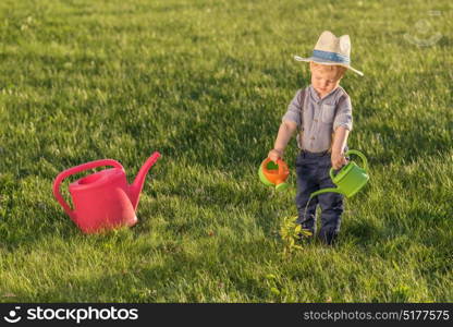 Portrait of toddler child outdoors. Rural scene with one year old baby boy wearing straw hat using watering can