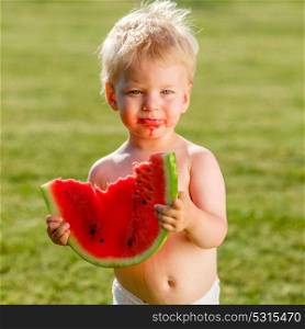 Portrait of toddler child outdoors. Rural scene with one year old baby boy eating watermelon slice in the garden. Dirty messy face of happy kid.