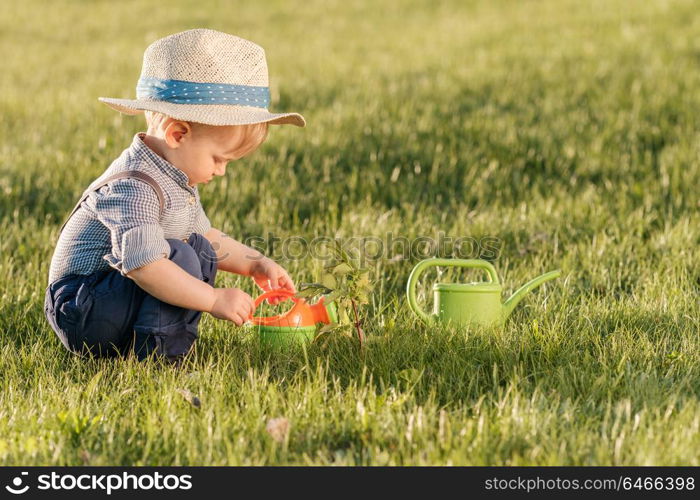 Portrait of toddler child outdoors. Rural scene with one year old baby boy wearing straw hat using watering can