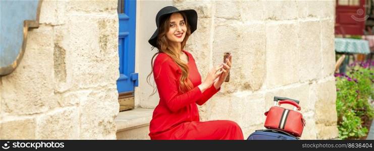 Portrait of tourist young caucasian woman in a red long dress and black hat with suitcase sitting on the stairs outdoors. Portrait of tourist young caucasian woman in a red long dress and black hat with suitcase sitting on the stairs outdoors.