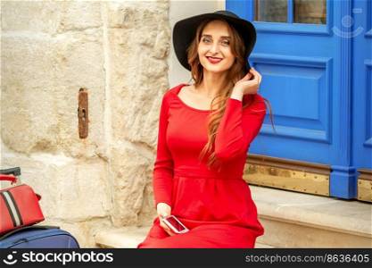 Portrait of tourist young caucasian woman in a red long dress and black hat with suitcase sitting on the stairs outdoors. Portrait of tourist young caucasian woman in a red long dress and black hat with suitcase sitting on the stairs outdoors.