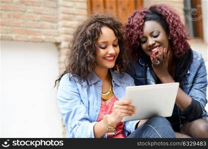 Portrait of two beautiful girls with tablet computer in urban backgrund, black and mixed women. Friends talking