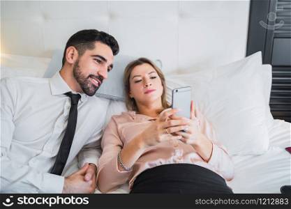 Portrait of two business people taking a break from work, lying on bed and using mobile phone at the hotel room. Business travel concept.