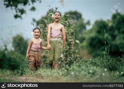 Portrait of two Cute girls in Thai traditional dress and put white flower on her ear walking hand in hand on rice field, They are smi≤with hapπ≠ss and looking at camera,©space
