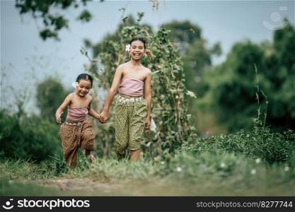 Portrait of two Cute girls in Thai traditional dress and put white flower on her ear walking hand in hand on rice field, They are smile with happiness and looking at camera, copy space