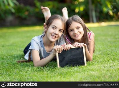 Portrait of two cute smiling girls lying on grass with chalkboard