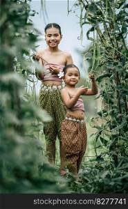 Portrait of two girls in Thai traditional dress and put white flower on her ear, standing in plant yard of long beans, They are smile with happiness and looking at camera
