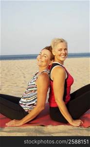 Portrait of two mature women sitting back to back on the beach and smiling