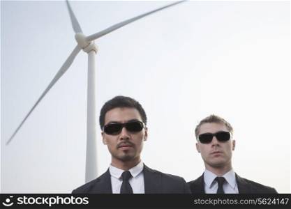 Portrait of two serious young businessmen in sunglasses standing by a wind turbine