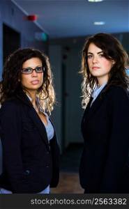 Portrait of two young businesswomen standing at office corridor, looking at camera.
