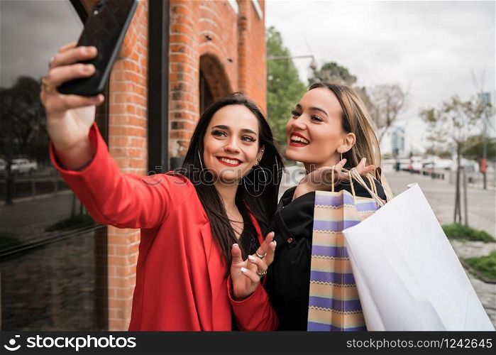 Portrait of two young friends enjoying doing shopping together while taking a selfie with phone at the street. Friendship and shopping concept.