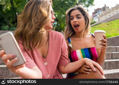 Portrait of two young friends enjoying together and using their mobile phone while sitting on stairs outdoors. Urban concept.