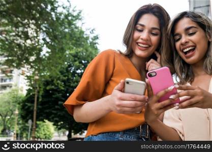 Portrait of two young friends enjoying together and using their mobile phones while standing outdoors. Urban concept.