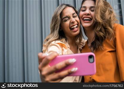 Portrait of two young friends having fun together and taking a selfie with a mobile phone outdoors. Urban concept.