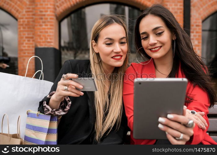 Portrait of two young friends shopping online with credit card and digital tablet while sitting outdoors. Friendship and lifestyle concept.
