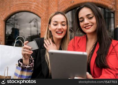 Portrait of two young friends shopping online with credit card and digital tablet while sitting outdoors. Friendship and lifestyle concept.