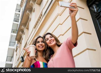 Portrait of two young friends smiling and taking a selfie with their mobile phone while sitting outdoors. Urban concept.