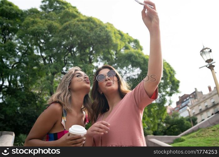 Portrait of two young friends smiling and taking a selfie with their mobile phone while standing outdoors. Urban concept.. Two friends taking selfie with phone outdoors.