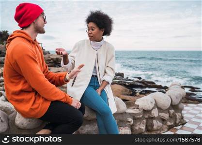 Portrait of two young friends spending good time together and having a conversation while sitting with the sea in the background.