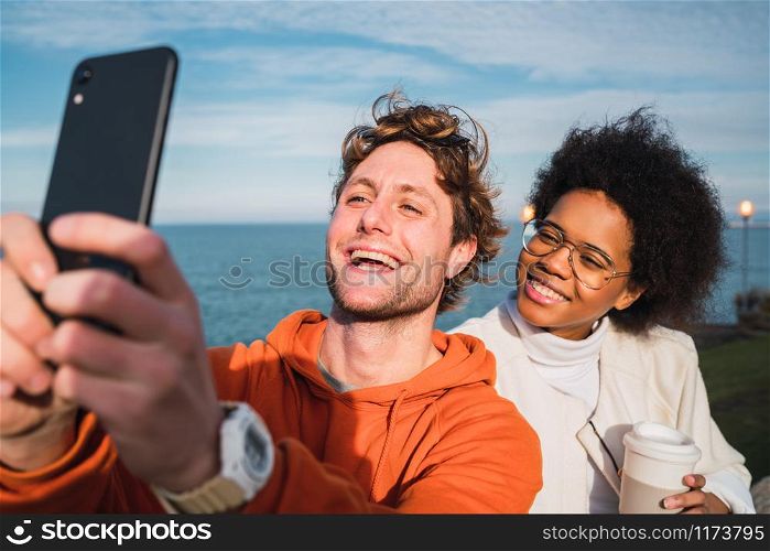 Portrait of two young friends spending good time together and taking a selfie with smartphone outdoors against the sea.