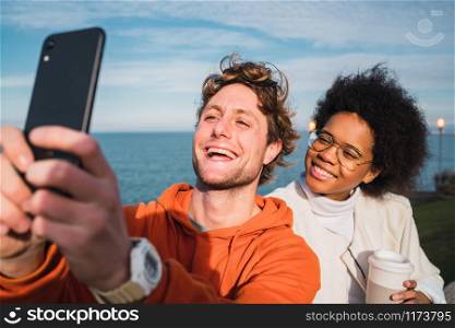 Portrait of two young friends spending good time together and taking a selfie with smartphone outdoors against the sea.