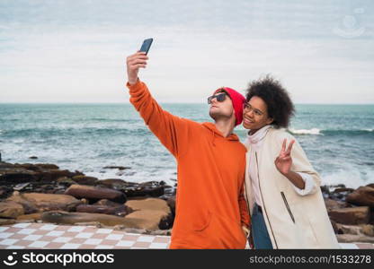 Portrait of two young friends spending good time together and taking a selfie with smartphone outdoors against the sea.