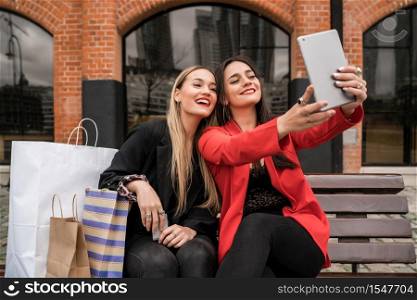 Portrait of two young friends taking a selfie with digital tablet while sitting outdoors with shopping bags. Friendship and lifestyle concept.