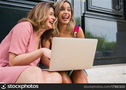 Portrait of two young friends using a laptop while sitting outdoors. Urban concept. Technology concept.