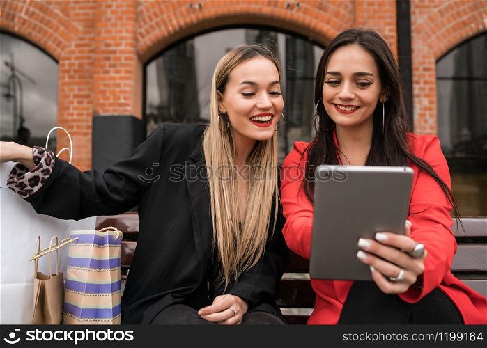 Portrait of two young friends using digital tablet while sitting outdoors with shopping bags. Friendship and lifestyle concept.