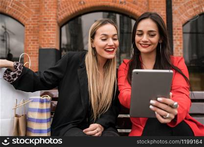 Portrait of two young friends using digital tablet while sitting outdoors with shopping bags. Friendship and lifestyle concept.