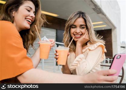 Portrait of two young friends using their mobile phone at a coffee shop outdoors. Urban concept.