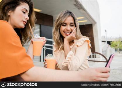 Portrait of two young friends using their mobile phone at a coffee shop outdoors. Urban concept.