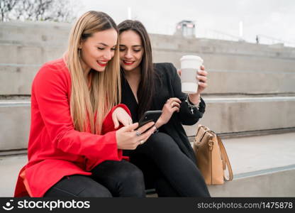 Portrait of two young friends using their mobile phone while sitting outdoors at the street. Lifestyle and friendship concept.