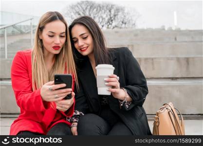 Portrait of two young friends using their mobile phone while sitting outdoors at the street. Lifestyle and friendship concept.