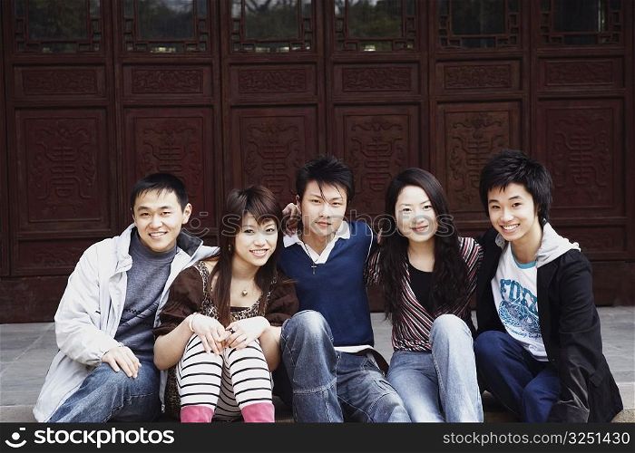 Portrait of two young women and three young men sitting in front of a wooden door