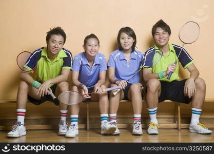 Portrait of two young women and two young men sitting side by side on a bench and holding badminton rackets