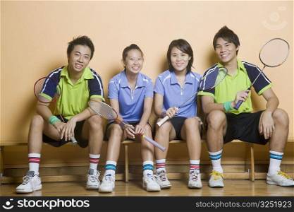 Portrait of two young women and two young men sitting side by side on a bench and holding badminton rackets