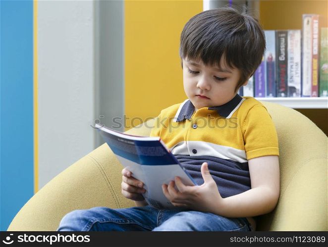 Portrait of unhappy kid sitting alone looking at the book with bored face in library, Upset chile boy with sad face with deep through sitting alone in book shop, Little boy with bored face