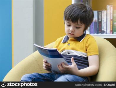 Portrait of unhappy kid sitting alone looking at the book with bored face in library, Upset chile boy with sad face with deep through sitting alone in book shop, Little boy with bored face